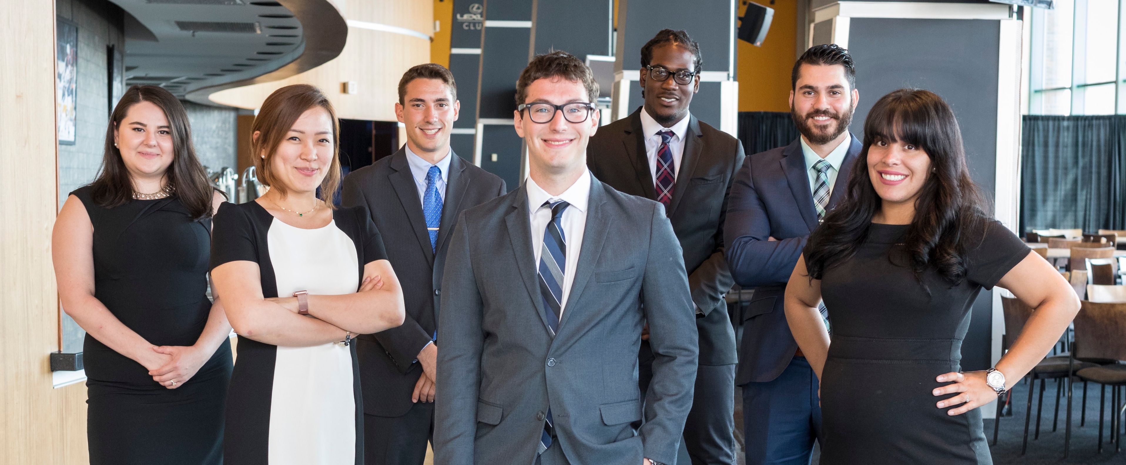 Seven MBA students dressed in professional business attire standing and looking at the camera. 